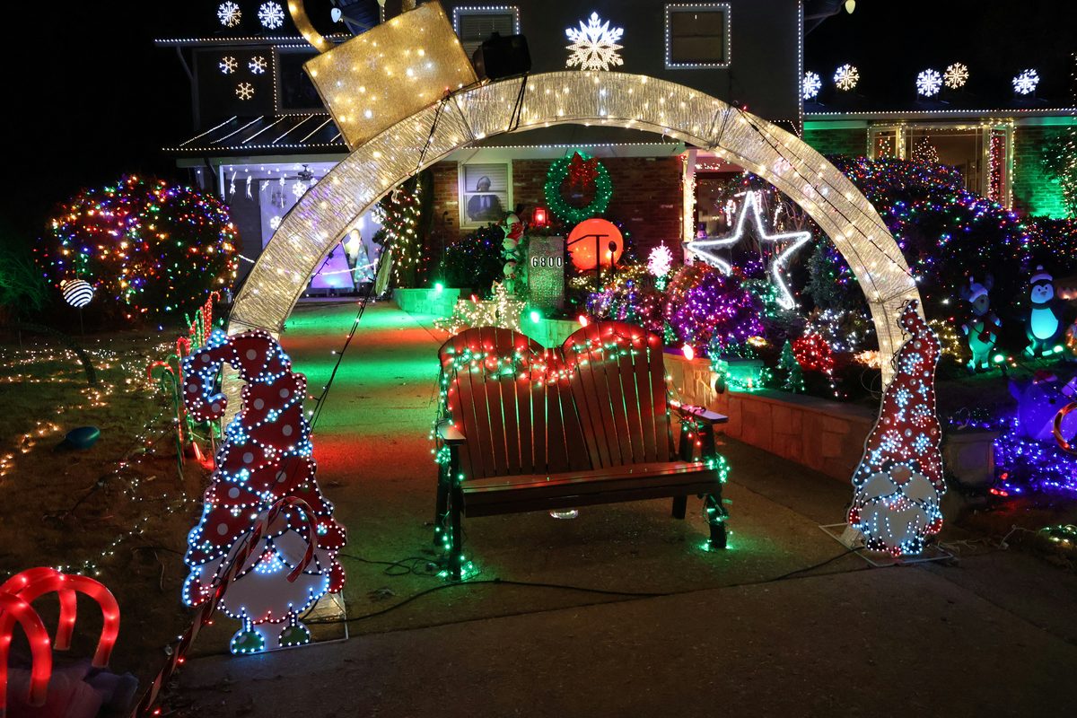 A front yard at night covered in Christmas lights, with a lit archway over the walk, a bench wrapped in lights and a large lit gnome