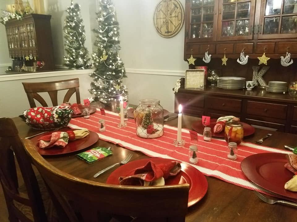 A dining table set for Christmas with a red striped runner, red chargers, festive napkins, candles and small trees in jars, with lit trees and a hutch hung with stars and doves behind