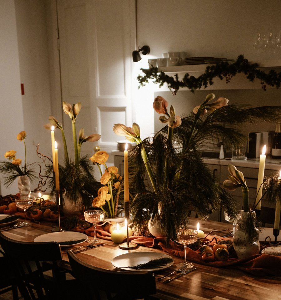 A dining table set for a seasonal dinner, with tall candles, floral and greenery arrangements, linens and a garland over the shelf