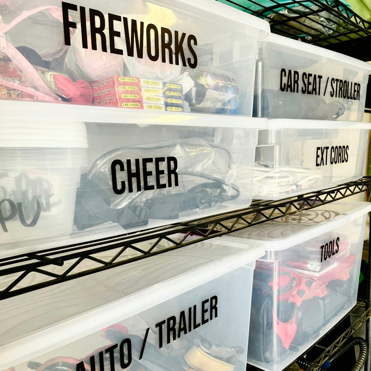 Clear plastic bins on wire garage shelving, each labeled in bold black letters: Fireworks, Car Seat / Stroller, Cheer, Ext Cords, Tools, Auto / Trailer